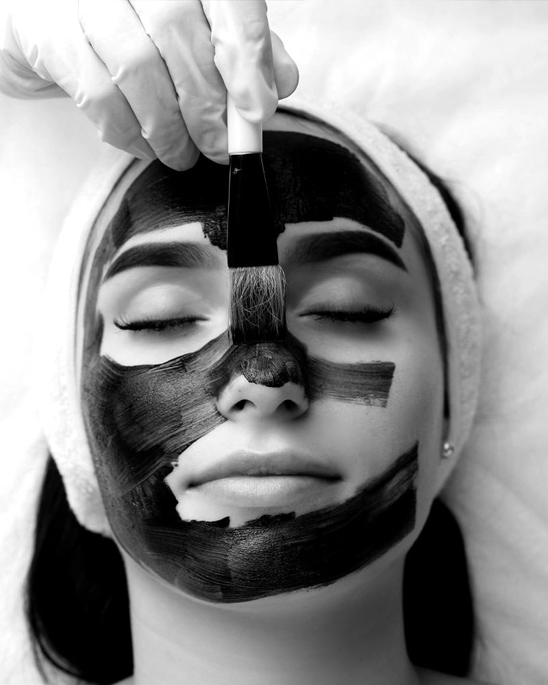 A woman with closed eyes receives a black facial mask, showcasing a relaxing facial in Nassau County, NY.