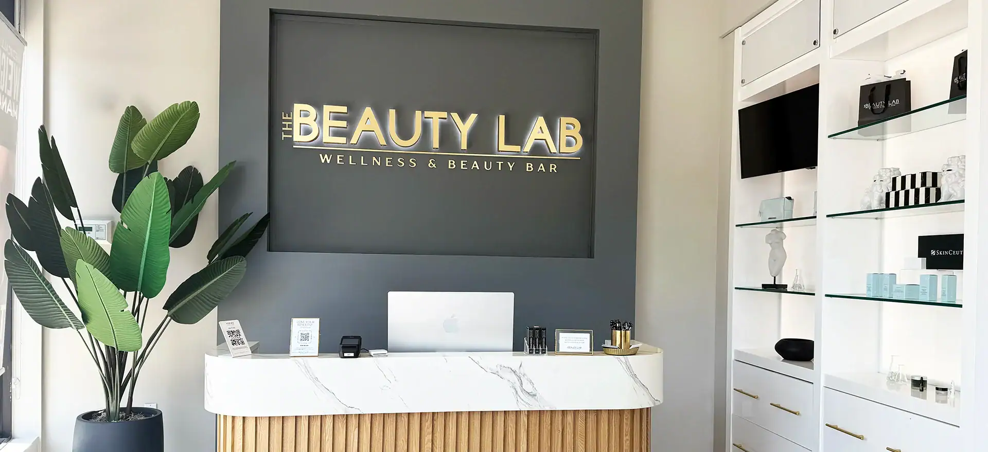 The Beauty Lab reception desk features a modern minimalist design, plants, and products displayed on shelves.