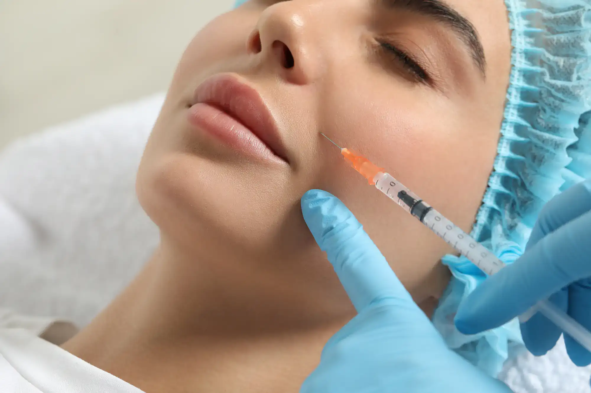 A woman lies down while a gloved medical professional gives her a cosmetic injection in the cheek.