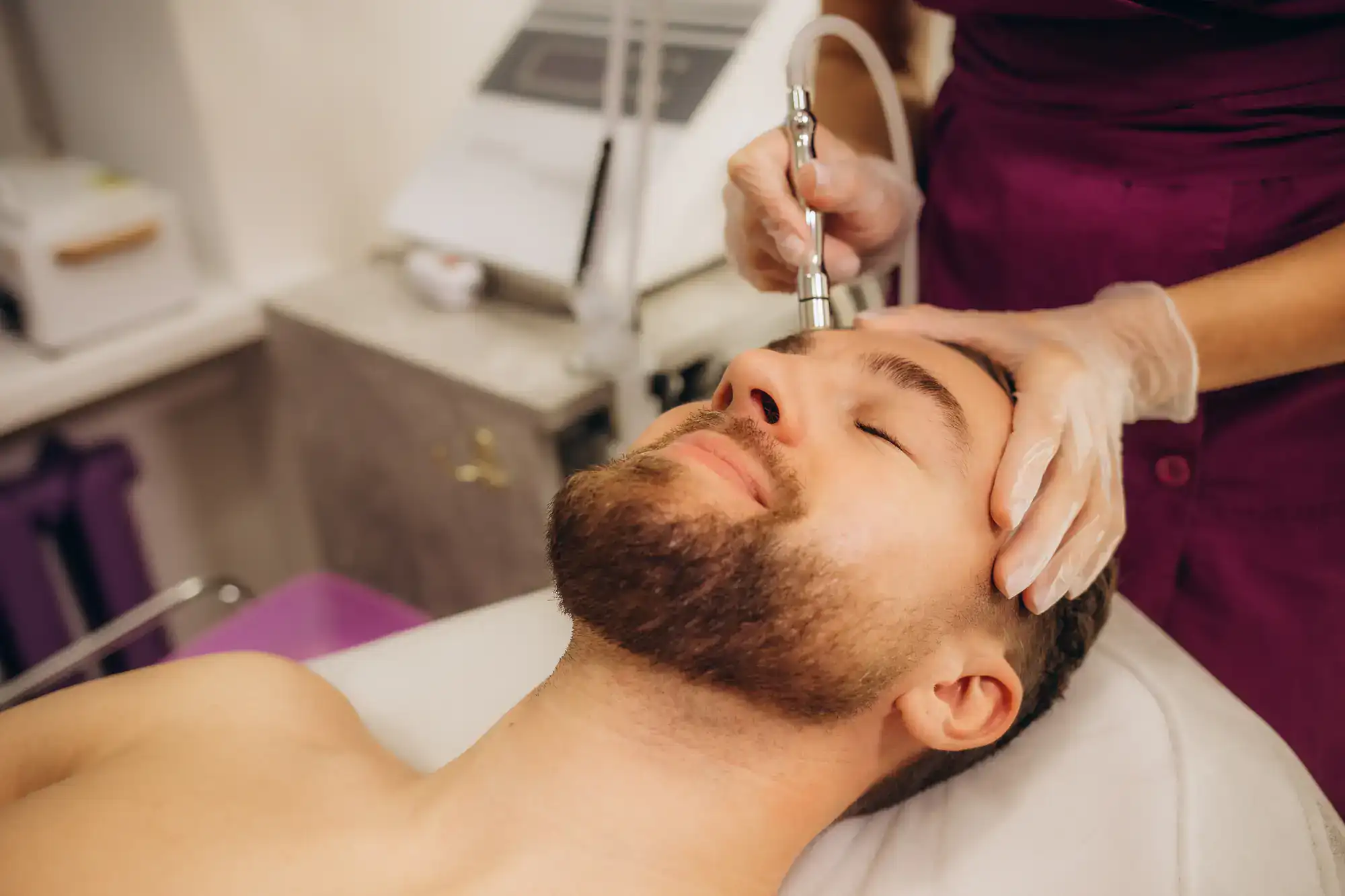A bearded man receives a facial treatment using a skincare device at a modern spa or clinic setting.