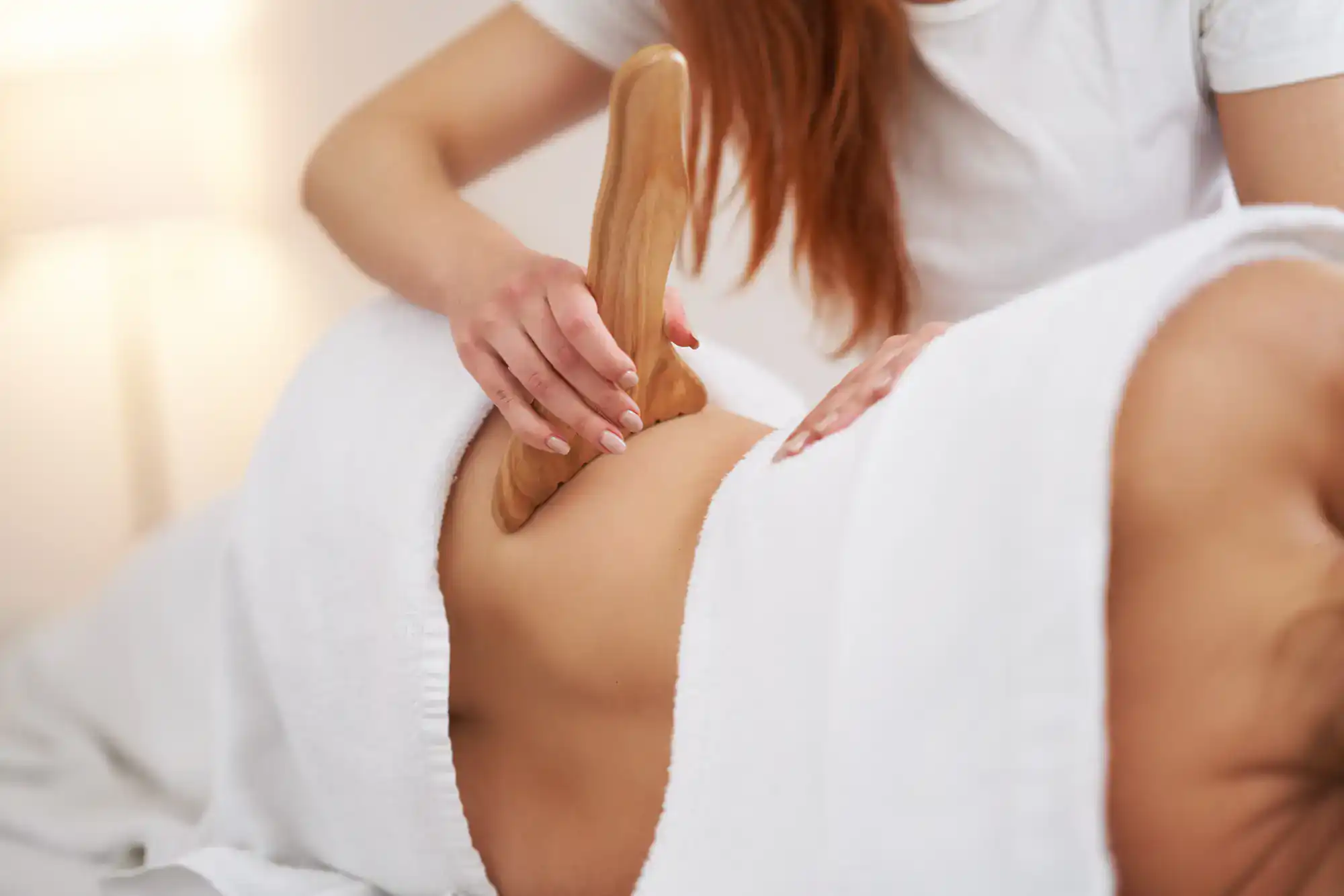 A person enjoys a wooden massage tool treatment on their back, covered by a white towel in a spa setting.