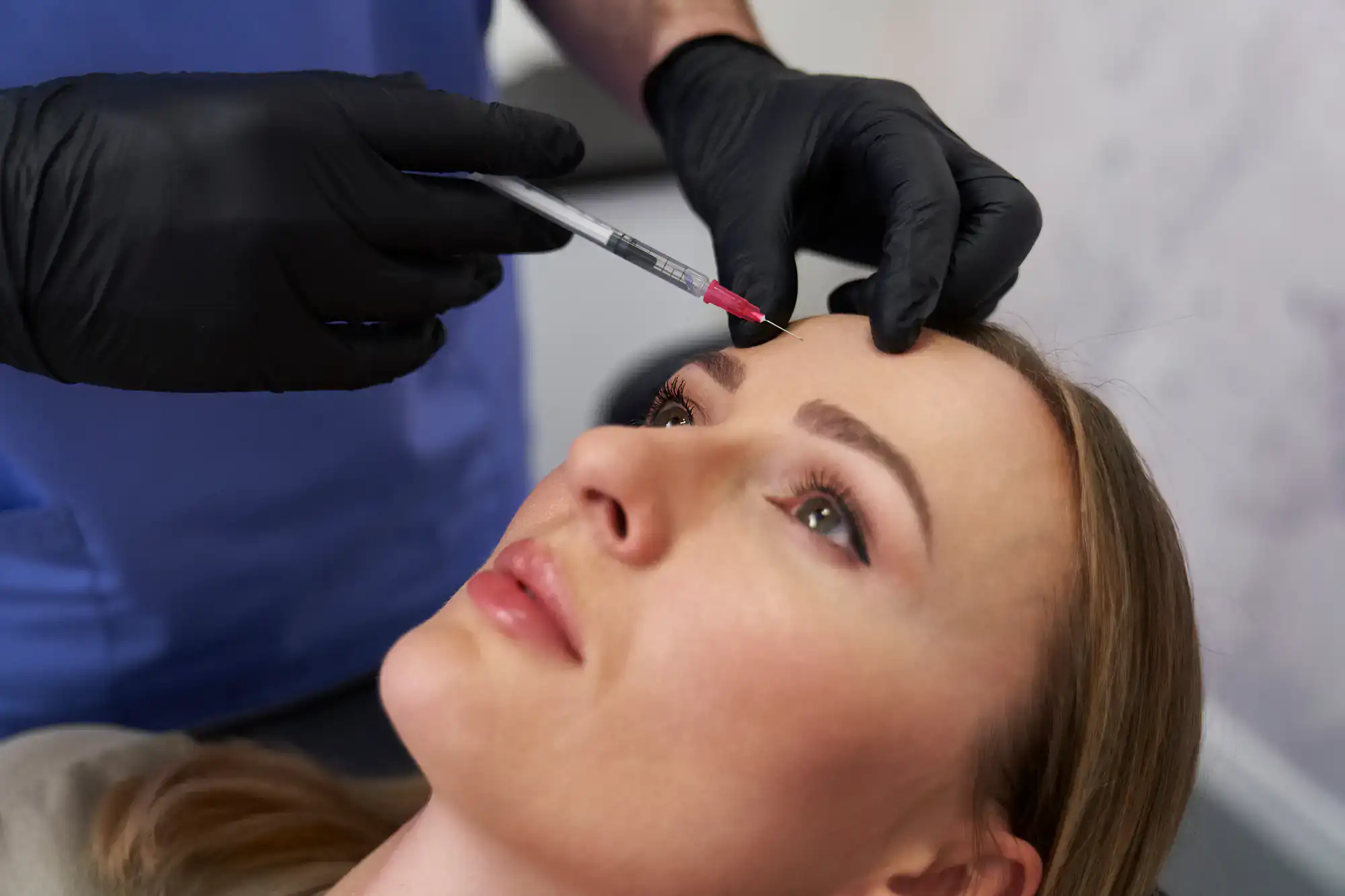 A woman receives a cosmetic injection in her forehead from a professional wearing black gloves.