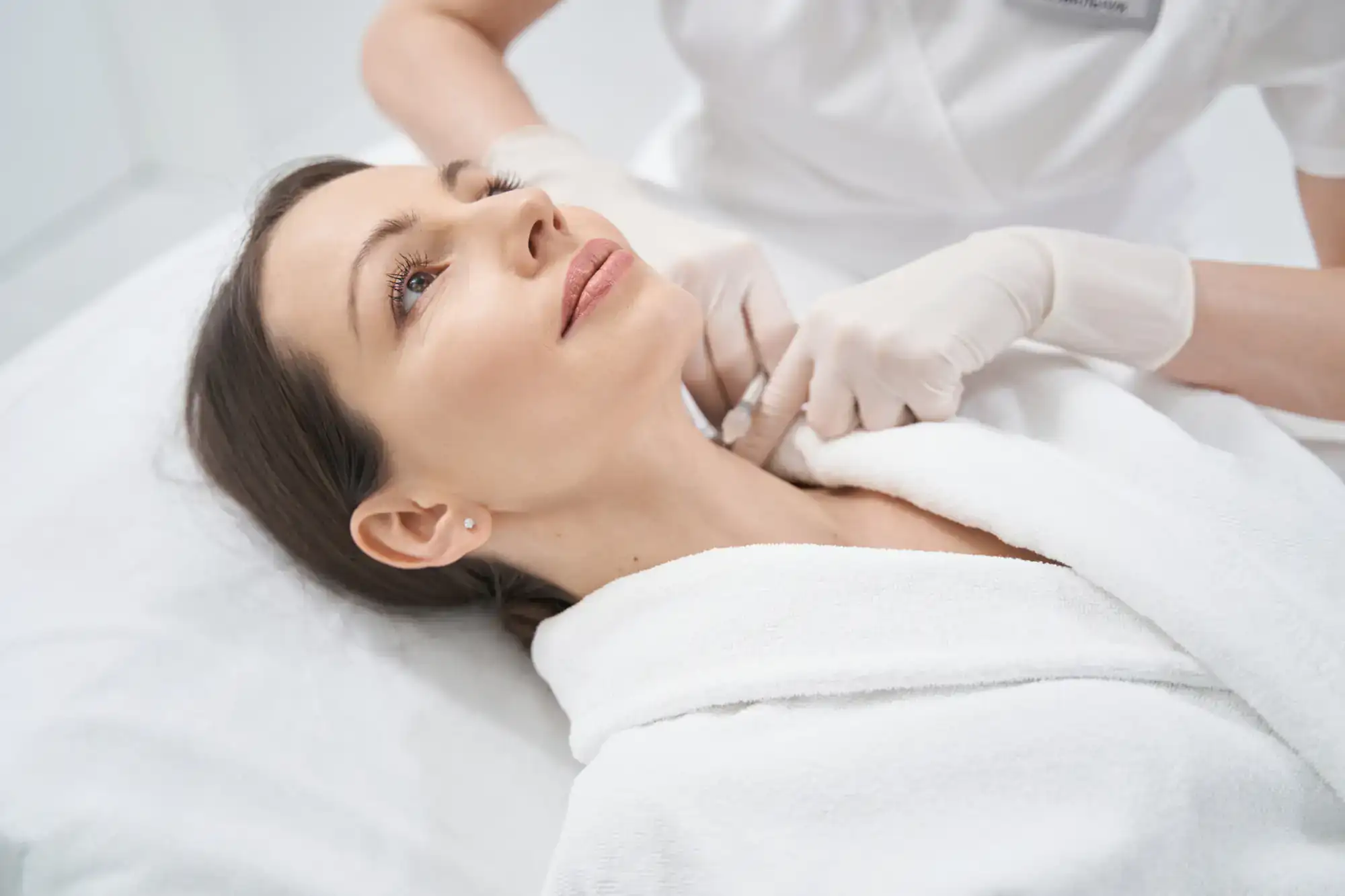 A woman in a white robe gets a neck cosmetic treatment from a professional wearing gloves.