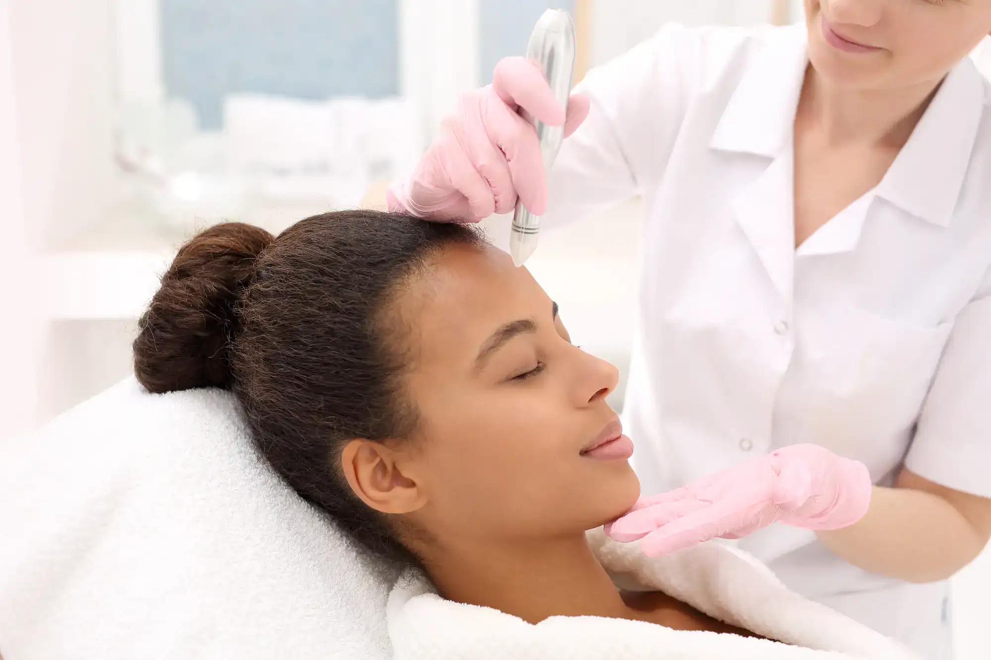 A woman receives a facial skin treatment from a professional in pink gloves at a relaxing spa setting.