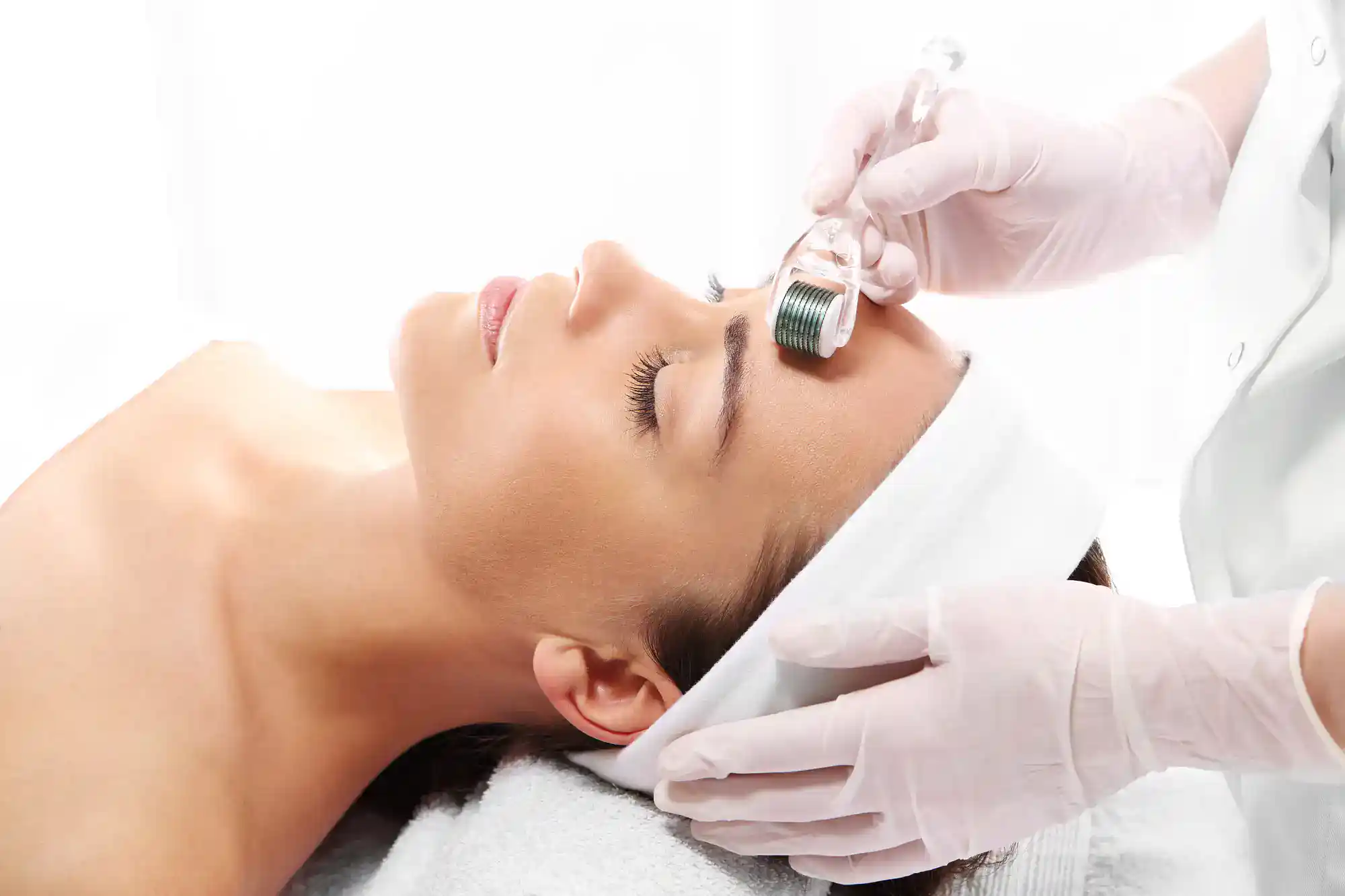A woman gets a microneedling facial as gloved hands use a dermaroller on her forehead at a skincare clinic.
