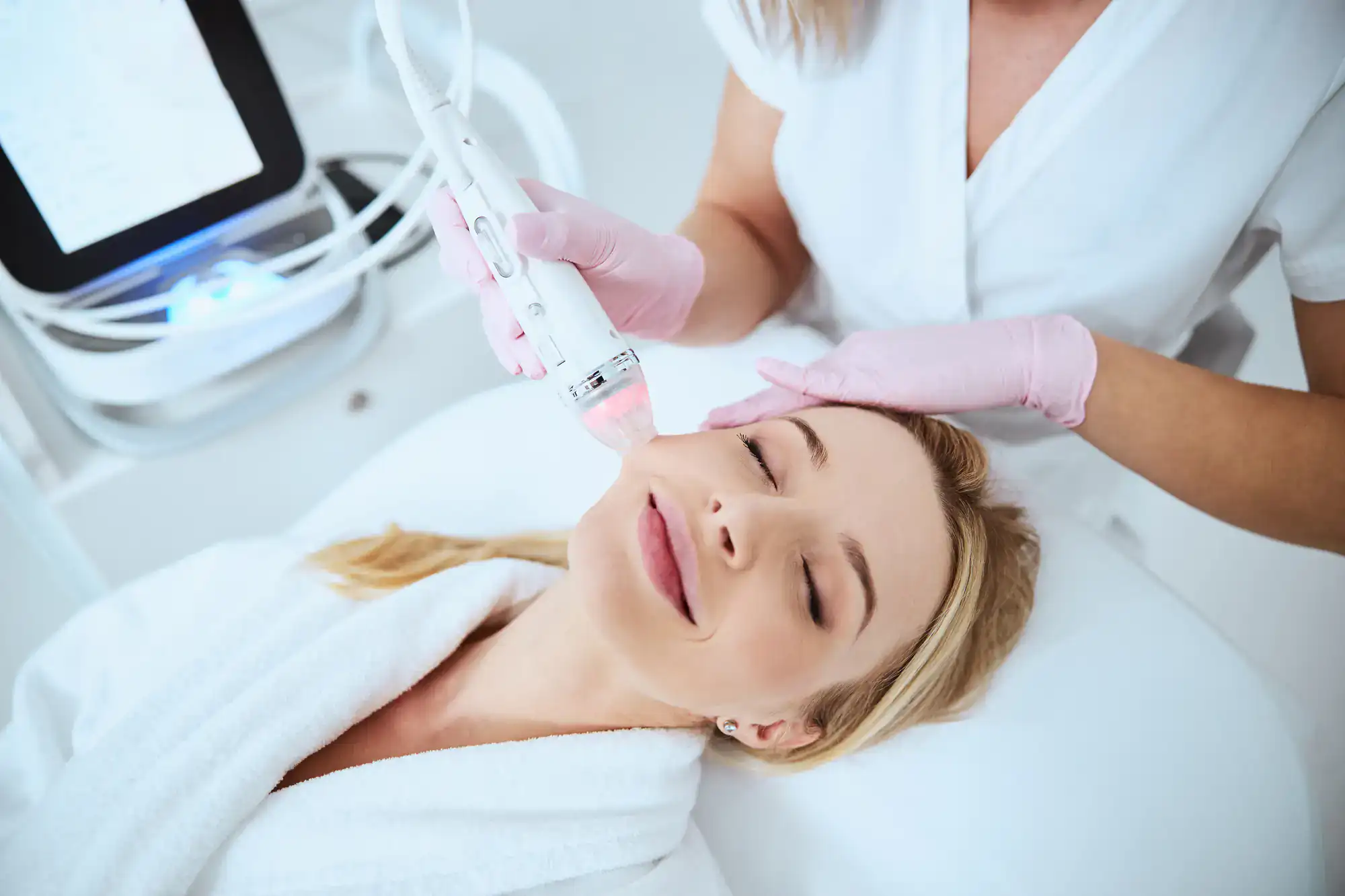 A woman in a white robe gets a facial skin treatment from a professional wearing pink gloves at a spa.