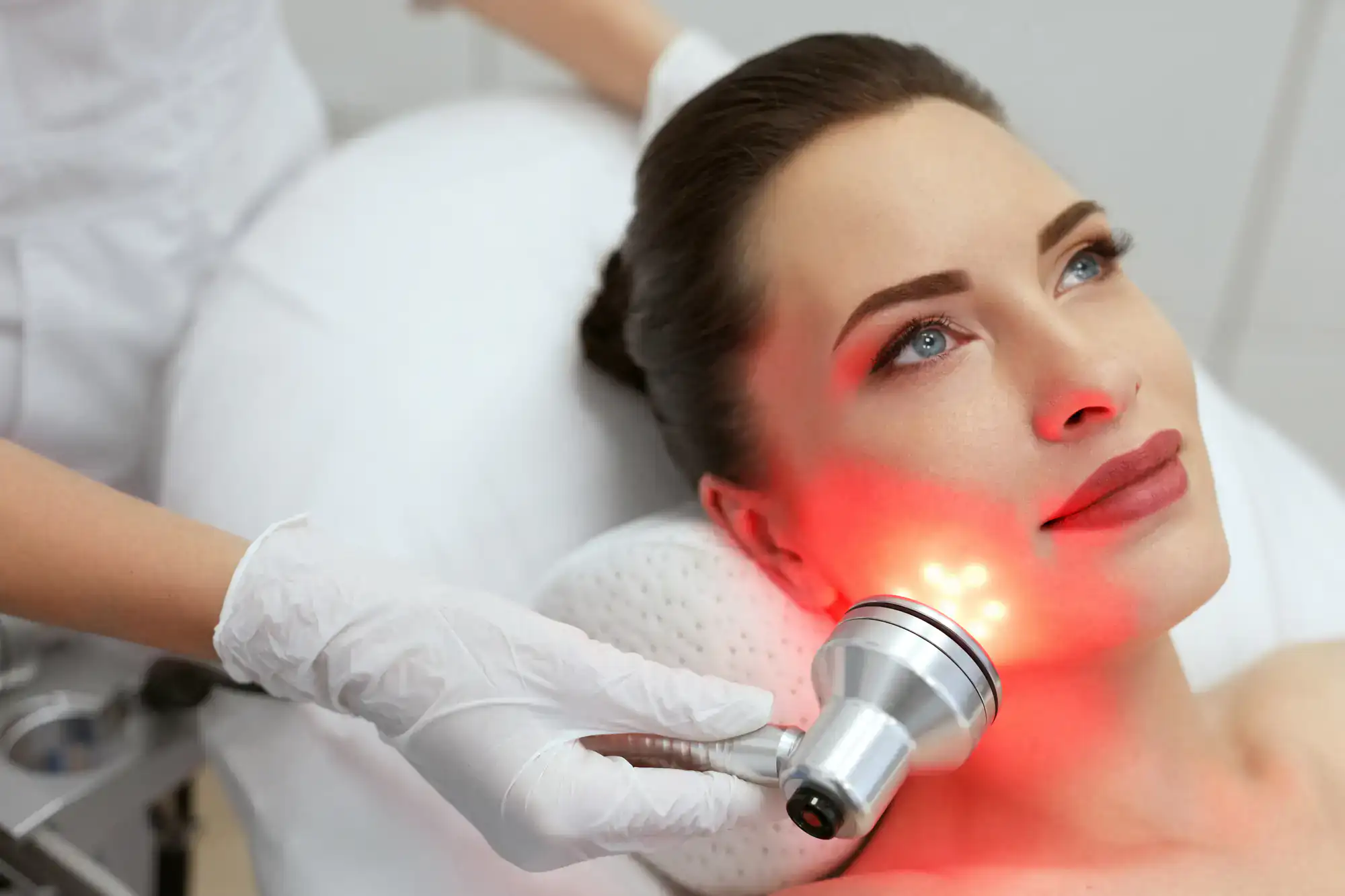 A woman receives red light therapy facial treatment from a professional in white gloves at a clinic.