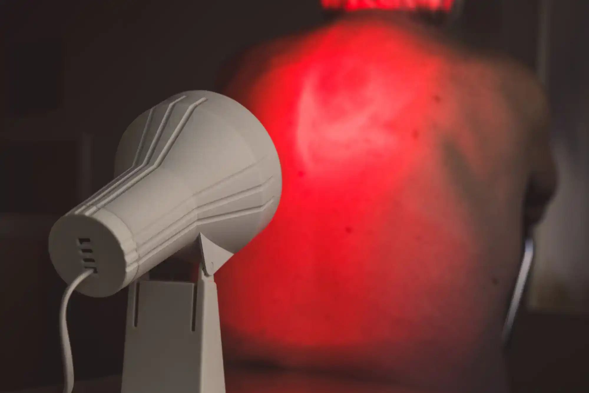 A person receives red light therapy on their bare back from a nearby lamp in a dimly lit room.