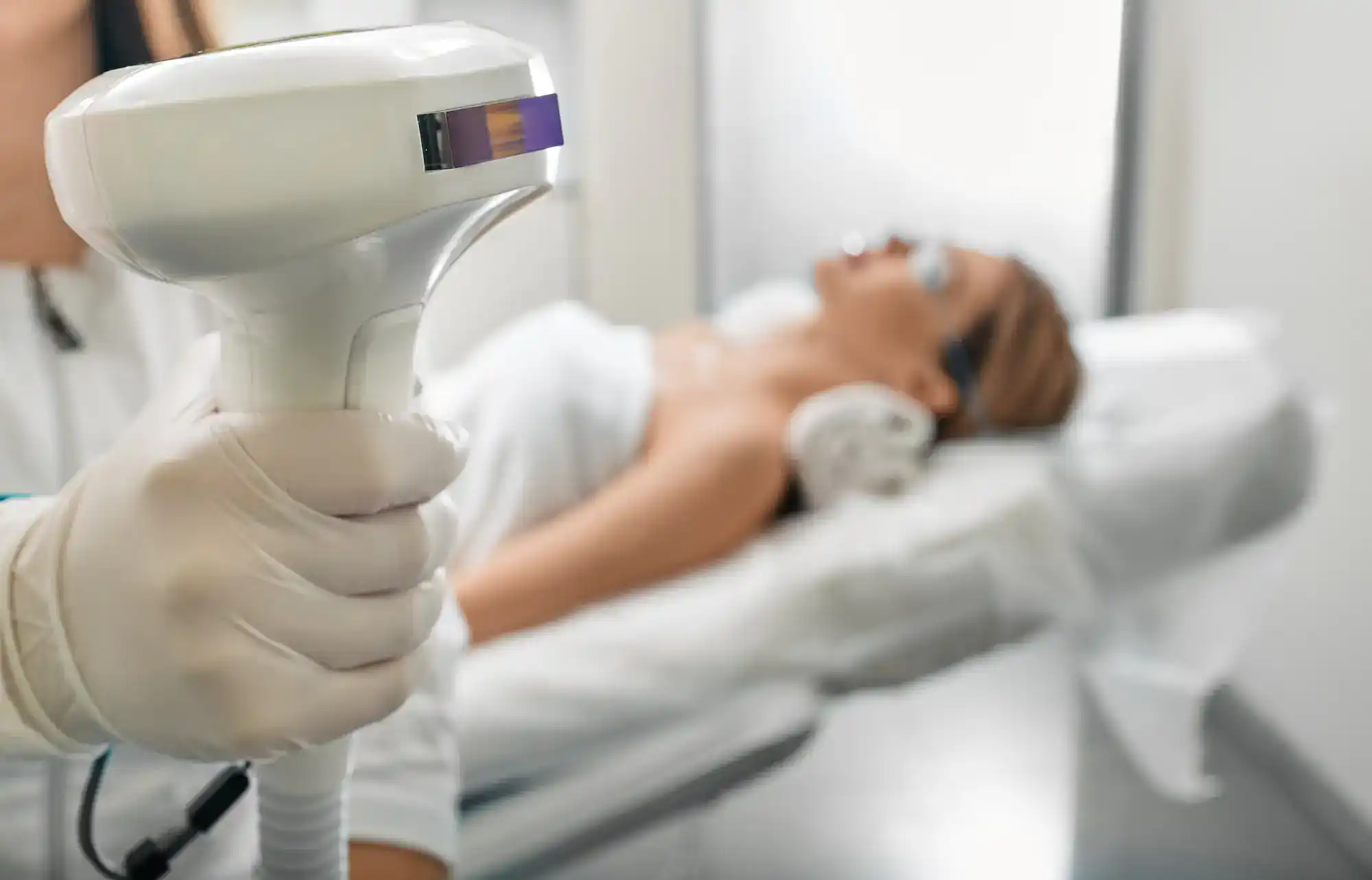 Aesthetic laser treatment shows a gloved hand using a laser on a woman with eye shields on a clinic bed.