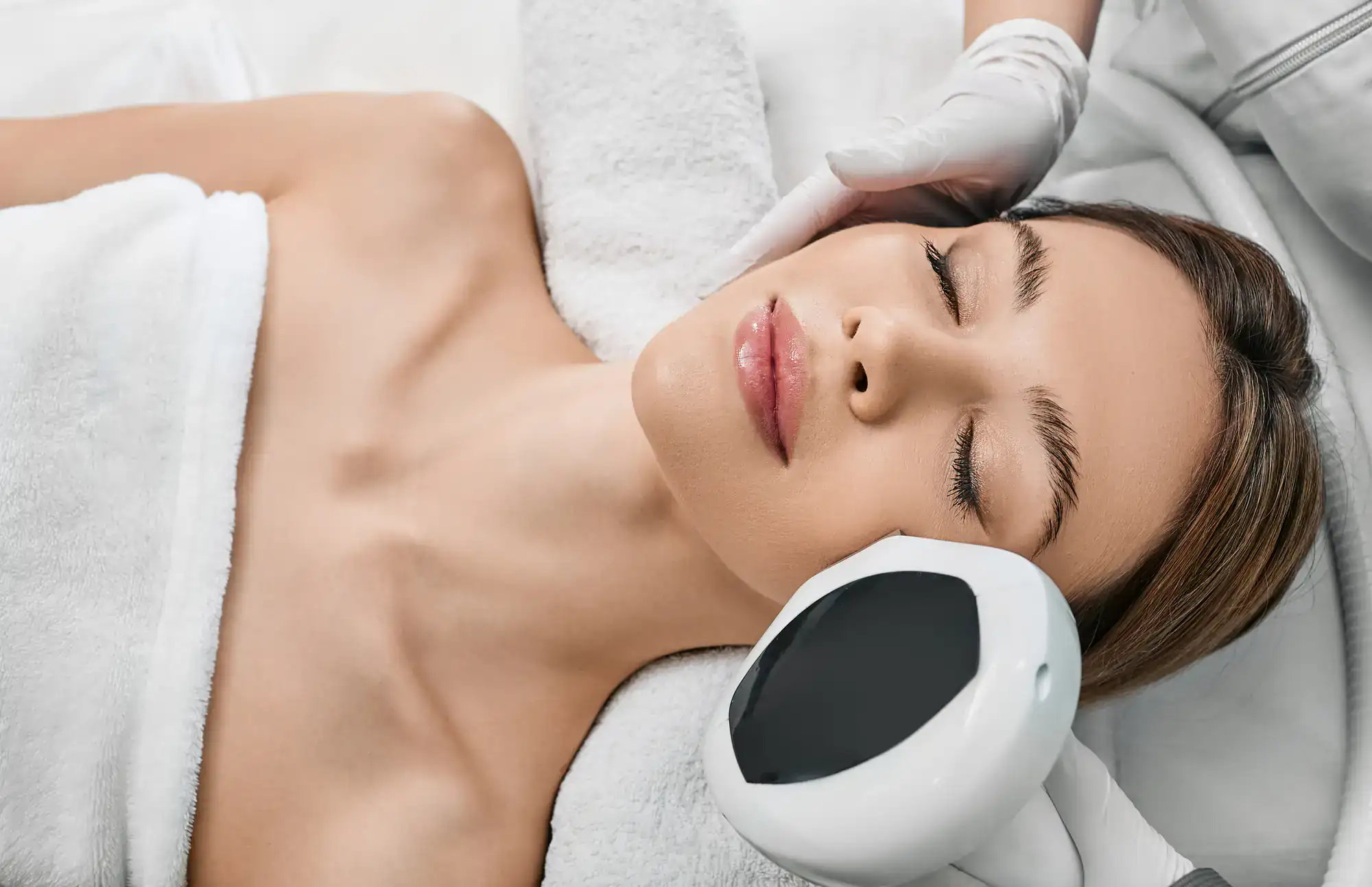 A woman lies with eyes closed, receiving a facial treatment from a gloved hand using a handheld device.