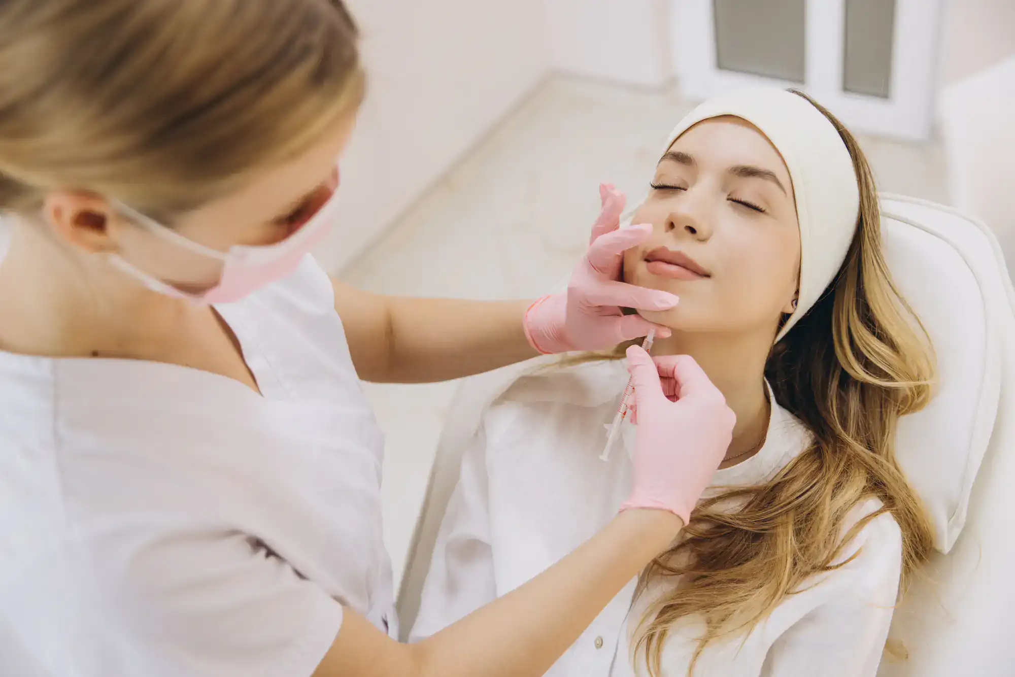 A woman receives a cosmetic injection in her face from a gloved, masked professional for facial treatment.