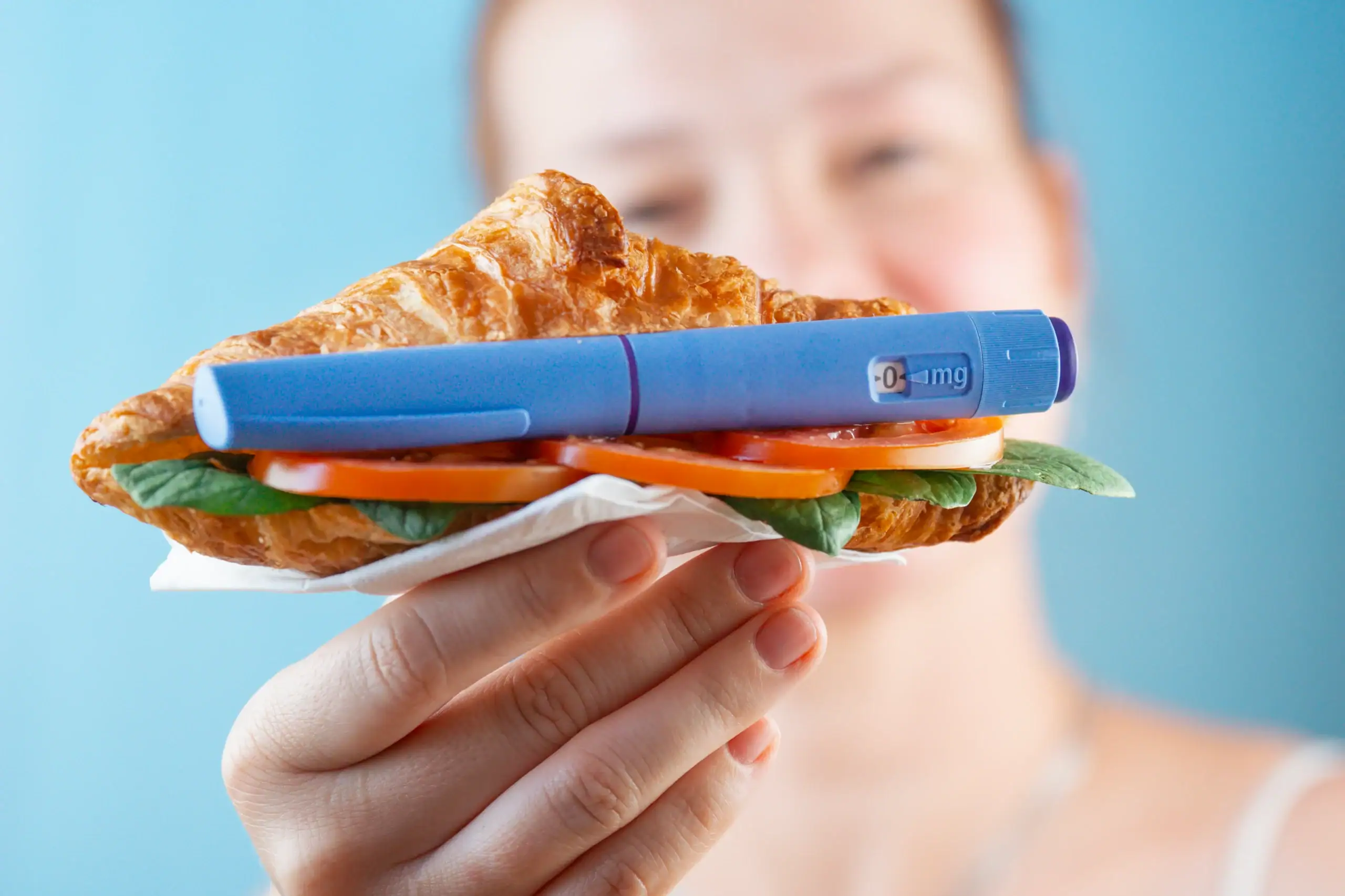 A person holds a croissant sandwich with a blue insulin pen on top, all set against a blue background.