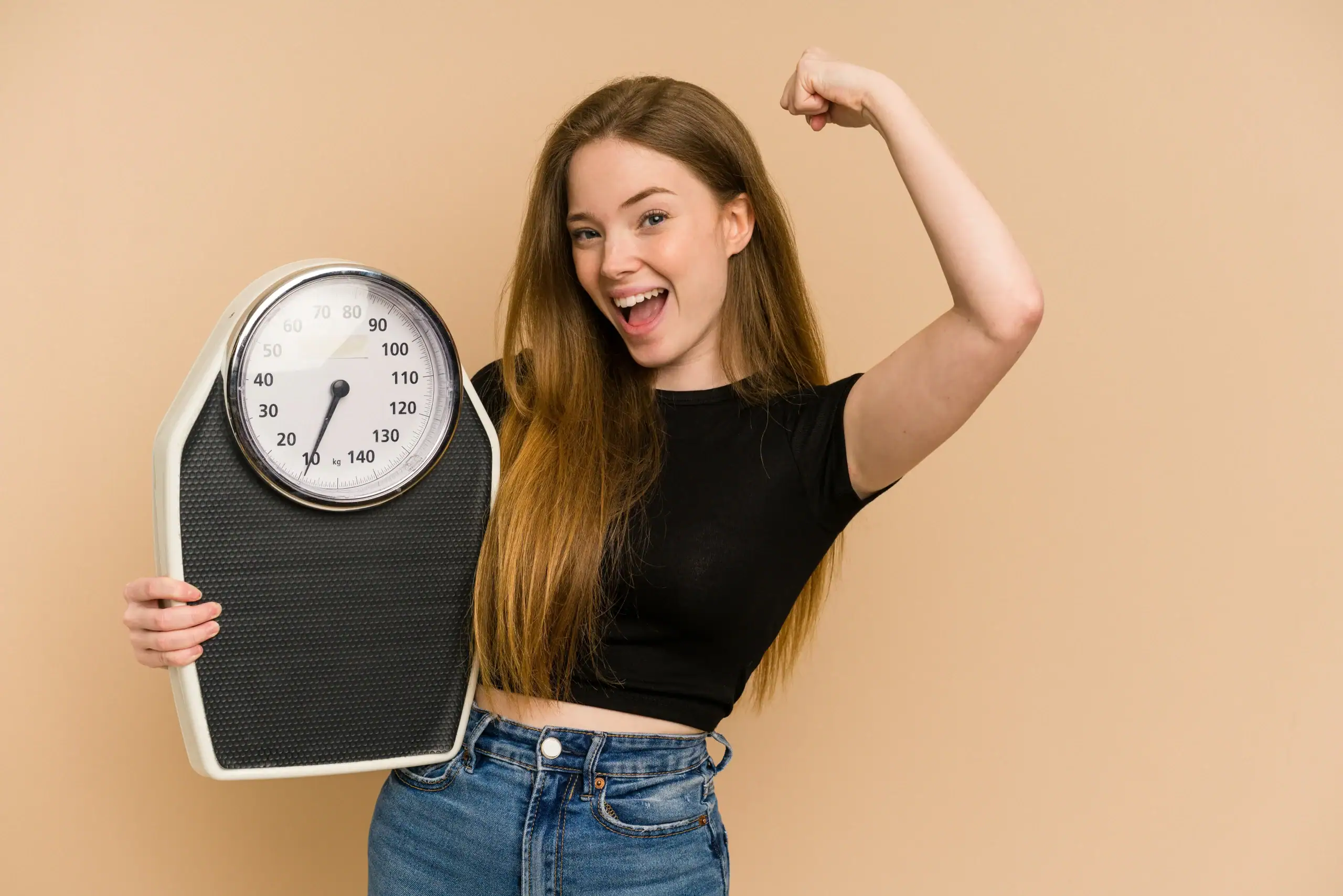 Smiling woman holds a scale and flexes her arm in a strength pose against neutral beige background.