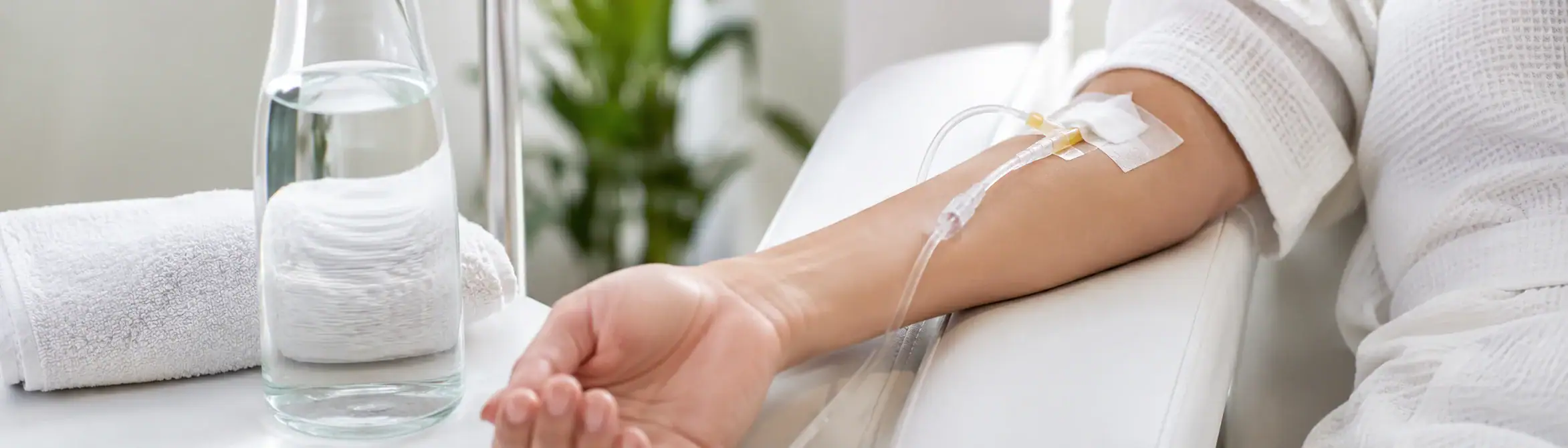 A person is receiving IV therapy in their arm while resting on a chair with a glass of water and towel nearby.