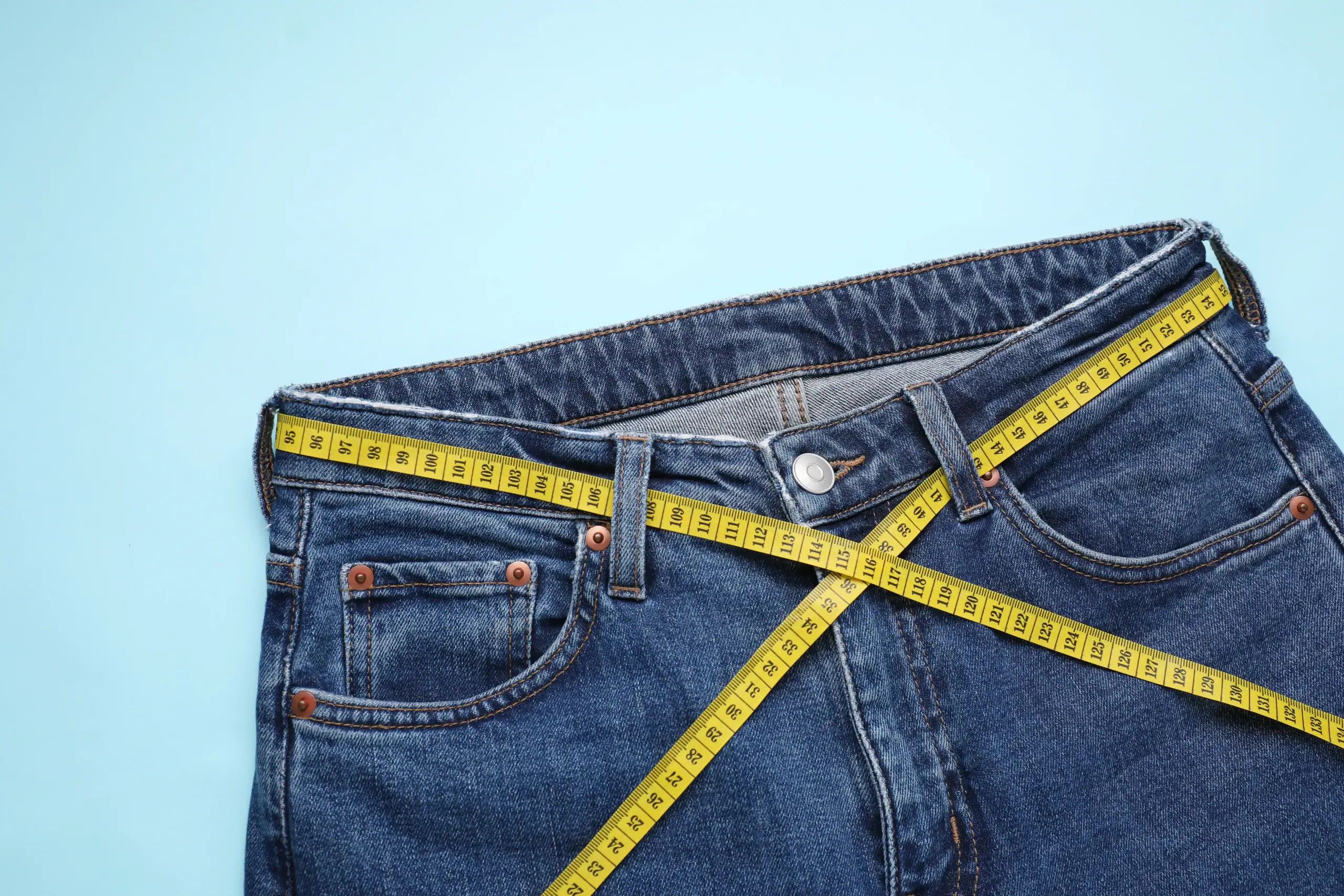 Blue jeans with a yellow measuring tape around the waist, displayed on a light blue background.