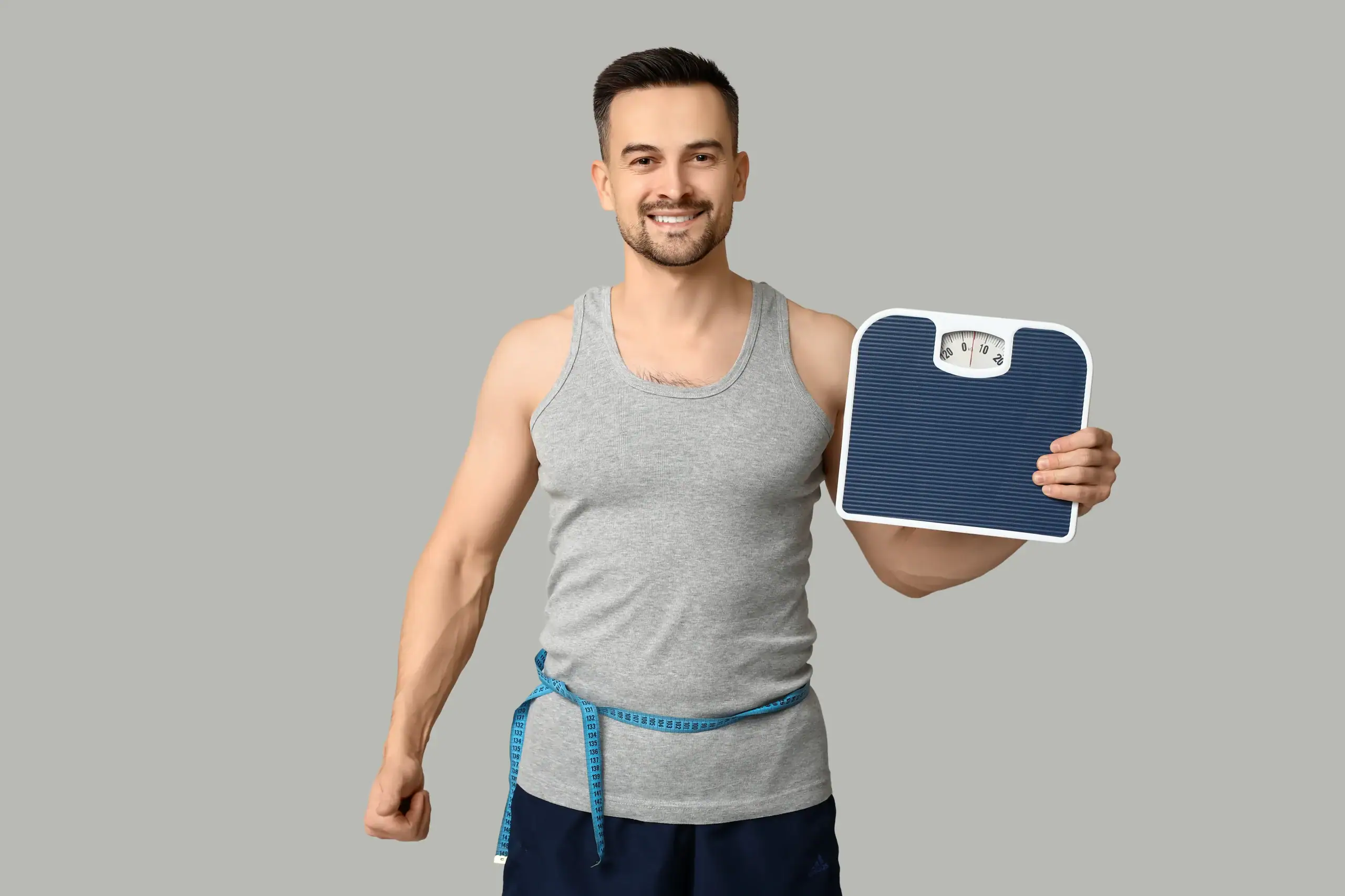 Smiling man in a gray tank top holds a bathroom scale and measures his waist with a blue tape.