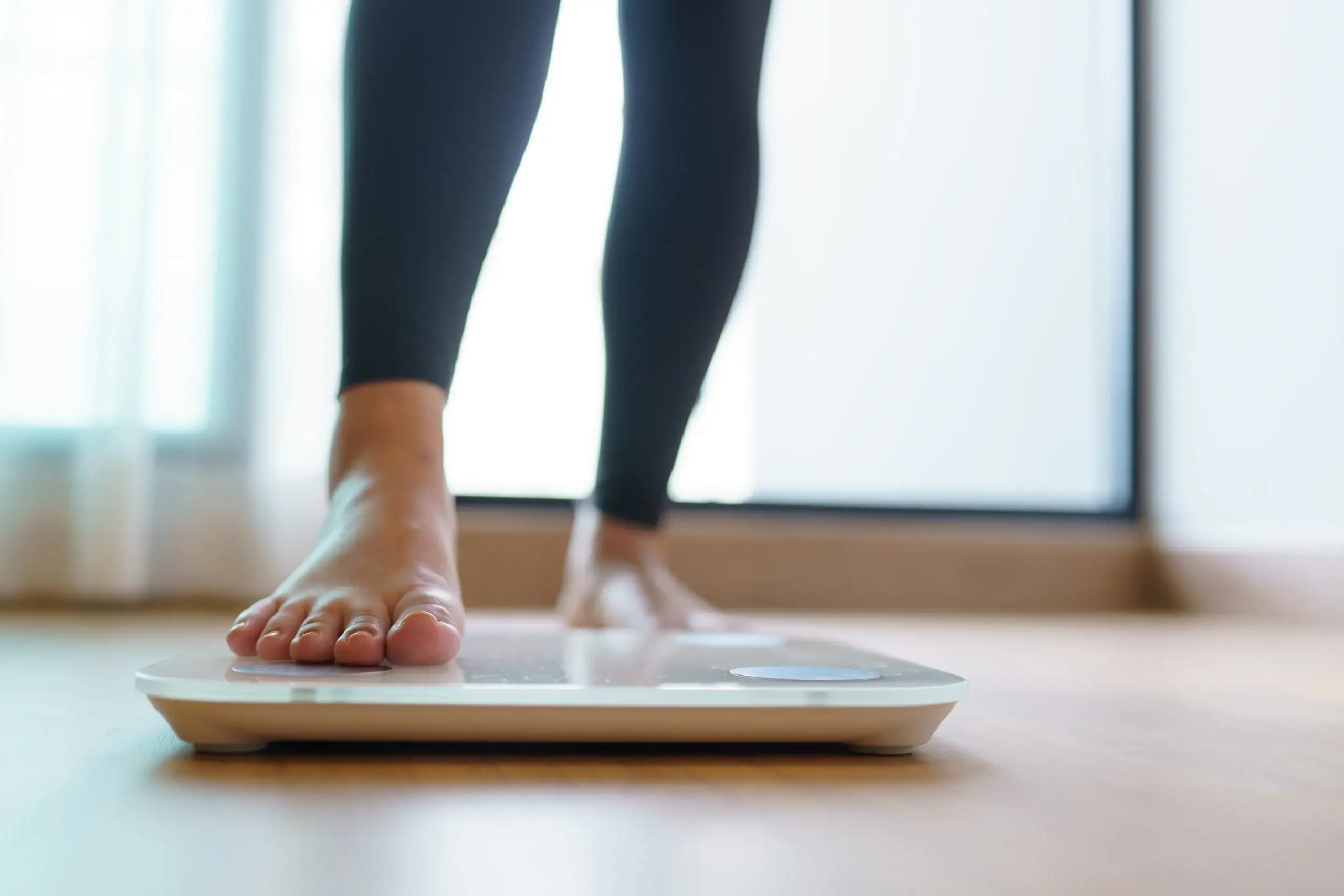 A person's bare feet step onto a digital bathroom scale on a wooden floor in an indoor setting.
