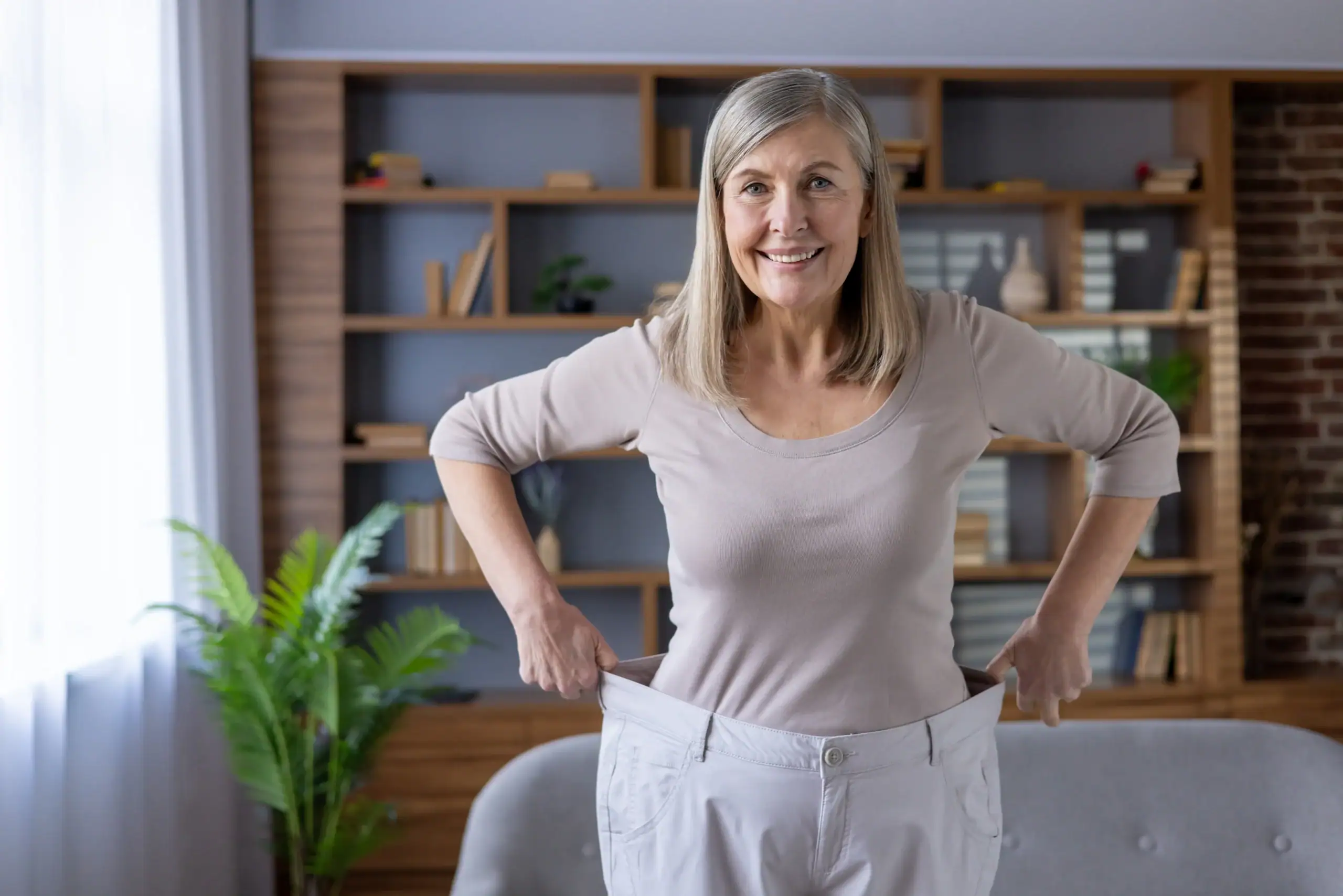 An older woman smiles indoors while holding up oversized pants to show impressive weight loss success.