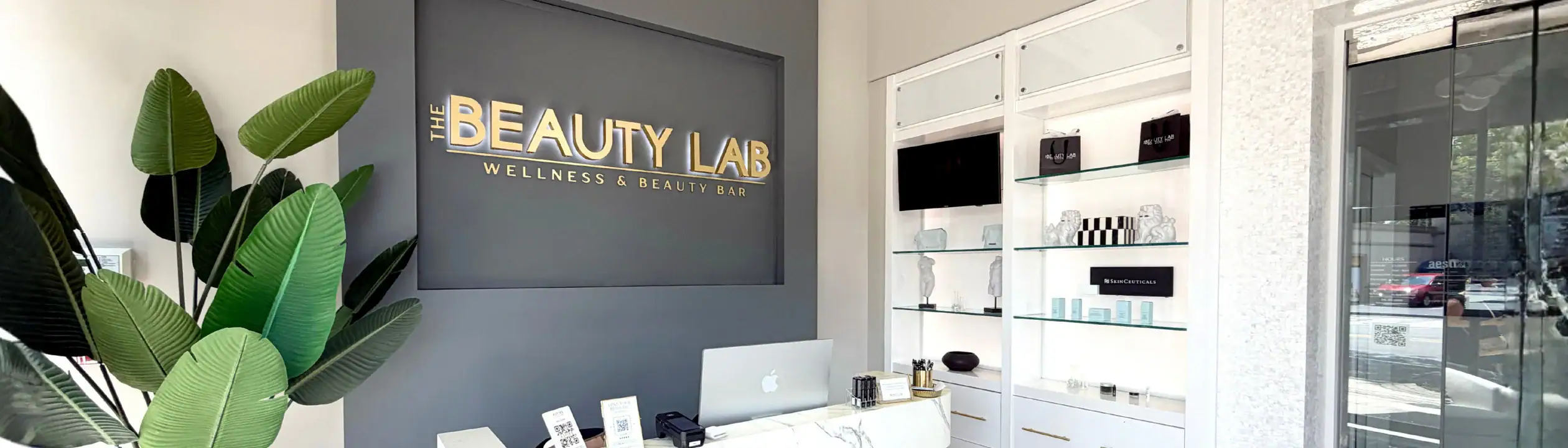 The Beauty Lab reception area features a front desk, wall sign, lush plants, and glass product shelves.