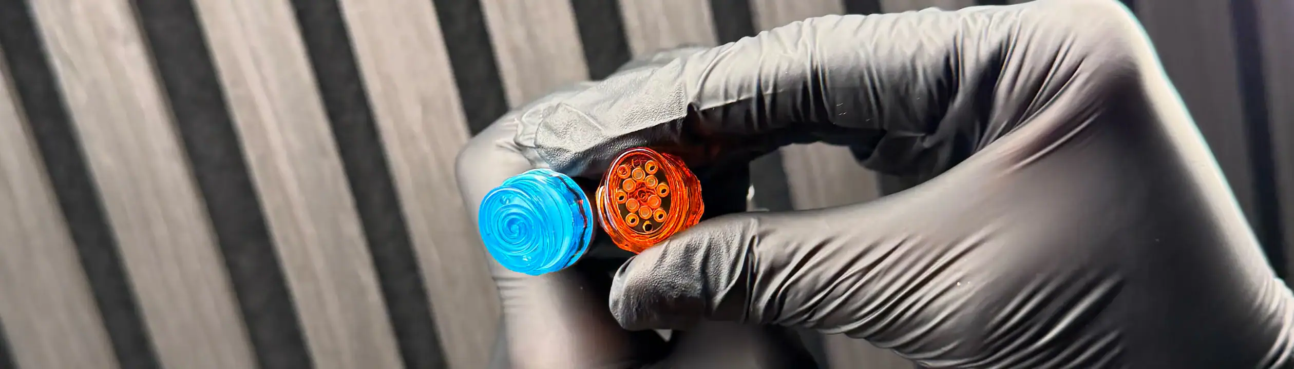 A gloved hand holds blue and orange circular electronic connectors against a striped background.