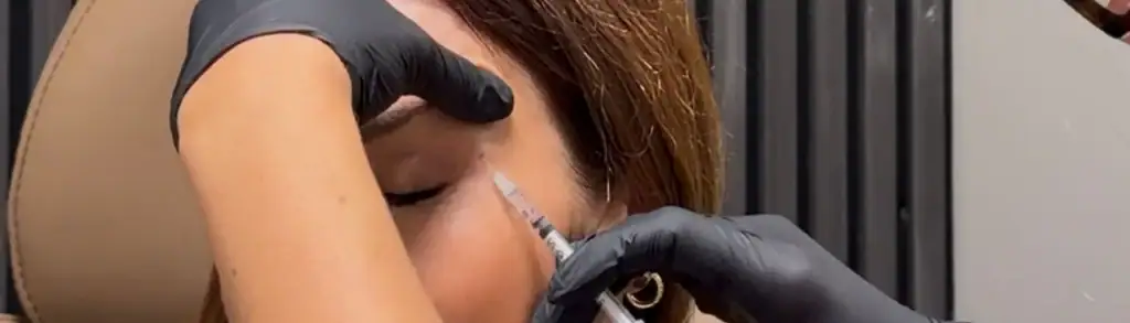 A person in a clinic with eyes closed receives a cosmetic injection near the eye from gloved hands.